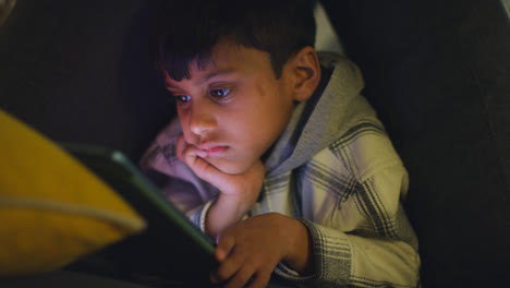 Close-Up-Of-Young-Boy-In-Home-Made-Camp-Made-From-Cushions-Playing-With-Digital-Tablet-At-Night-3